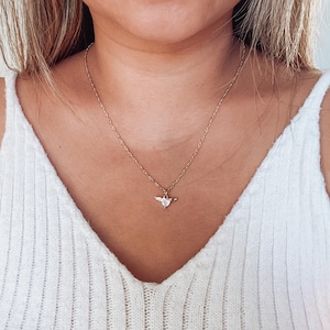 May include: A gold necklace with a fine chain and a pendant. The pendant has a clear, heart-shaped stone with wing details. The necklace is worn against a white vest top.