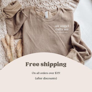 May include: A beige sweatshirt with the words "an angel calls me mama" in white script. The sweatshirt is folded on a textured surface with dried plants. The image also includes text that says "Free shipping".