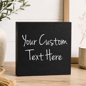 May include: A square black wooden block sign featuring the white text Your Custom Text Here in a handwritten script font. The sign is positioned on a wooden surface with the label Black/Font 1 printed in the bottom right corner.