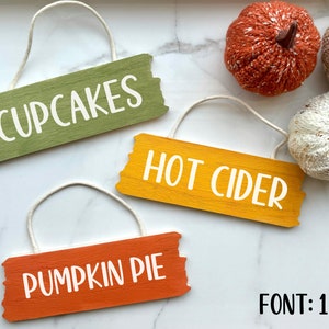 May include: Three wooden signs with white text on a white marble background. The signs are hanging from twine and read "CUPCAKES", "HOT CIDER", and "PUMPKIN PIE". The text is in a handwritten font. The signs are surrounded by knitted pumpkins.