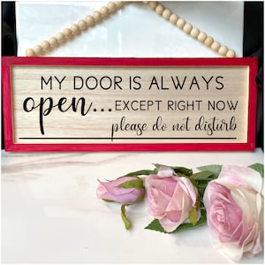 May include: A wooden sign with a red border and the text "My door is always open... except right now please do not disturb". The sign is hanging from a string of wooden beads.