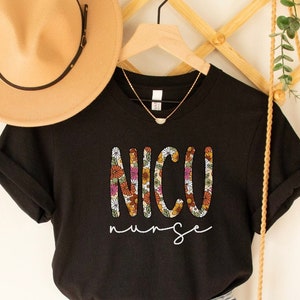 May include: Black t-shirt with the text "NICU nurse" in white with a floral pattern in the letters. The shirt is hanging on a wooden hanger with a brown hat and a straw bag.