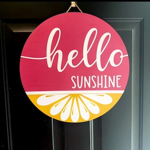 Könnte beinhalten: Runder Holzschild mit leuchtend pinkfarbenem Hintergrund und dem weißen Schriftzug "hello SUNSHINE". Die untere Hälfte zeigt ein gelb-weißes Zitrusfrucht-Design. Zum Aufhängen ist eine Juteschnur befestigt.