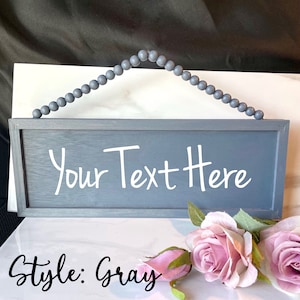 Peut inclure: Panneau rectangulaire en bois gris avec le texte "Your Text Here" en blanc. Le panneau est suspendu par une cha&icirc;ne de perles grises. Les mots "Style: Gray" sont &eacute;crits en script noir en bas. Des roses roses sont au premier plan.
