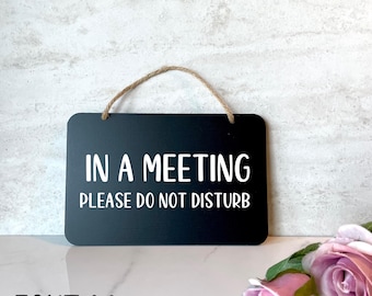 May include: Black sign with white text that reads "In a meeting, please do not disturb". The sign is hanging from a string.