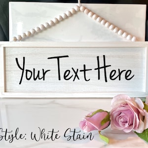 May include: A rectangular, white-stained wooden sign with the text "Your Text Here" in black. The sign is framed and has a string of wooden beads above it. Pink roses are in the foreground.
