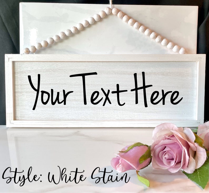May include: A rectangular, white-stained wooden sign with the text "Your Text Here" in black. The sign is framed and has a string of wooden beads above it. Pink roses are in the foreground.