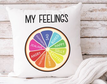May include: White square pillow with the text "MY FEELINGS" in black. The pillow features a colorful graphic of a citrus fruit slice divided into segments, each labeled with a different emotion, such as "Happy" and "Sad".