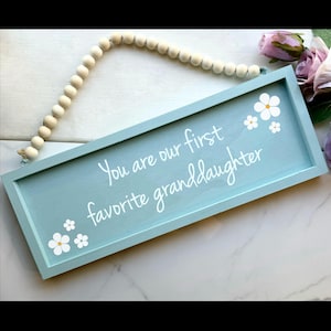 May include: A rectangular, light blue wooden sign with the text "You are our first favorite granddaughter" in white script. White flower decals are in the corners. The sign is suspended by a beaded wooden strand.