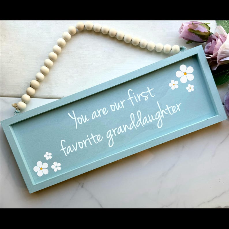 May include: A rectangular, light blue wooden sign with the text "You are our first favorite granddaughter" in white script. White flower decals are in the corners. The sign is suspended by a beaded wooden strand.