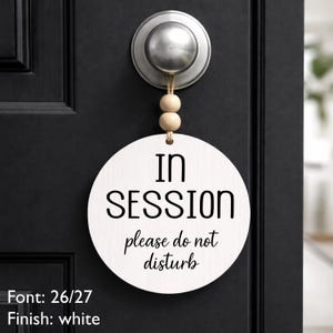 May include: A round white wooden sign hanging from a door handle by a jute string with two light wood beads. The sign features black text that reads IN SESSION and please do not disturb. The text Font: 26/27 and Finish: white are visible.