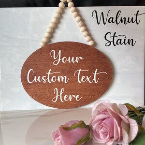 Pu&ograve; includere: Un cartello ovale in legno con una macchia di noce, appeso a una corda di perline bianche. Il cartello ha un testo bianco che dice "Your Custom Text Here".