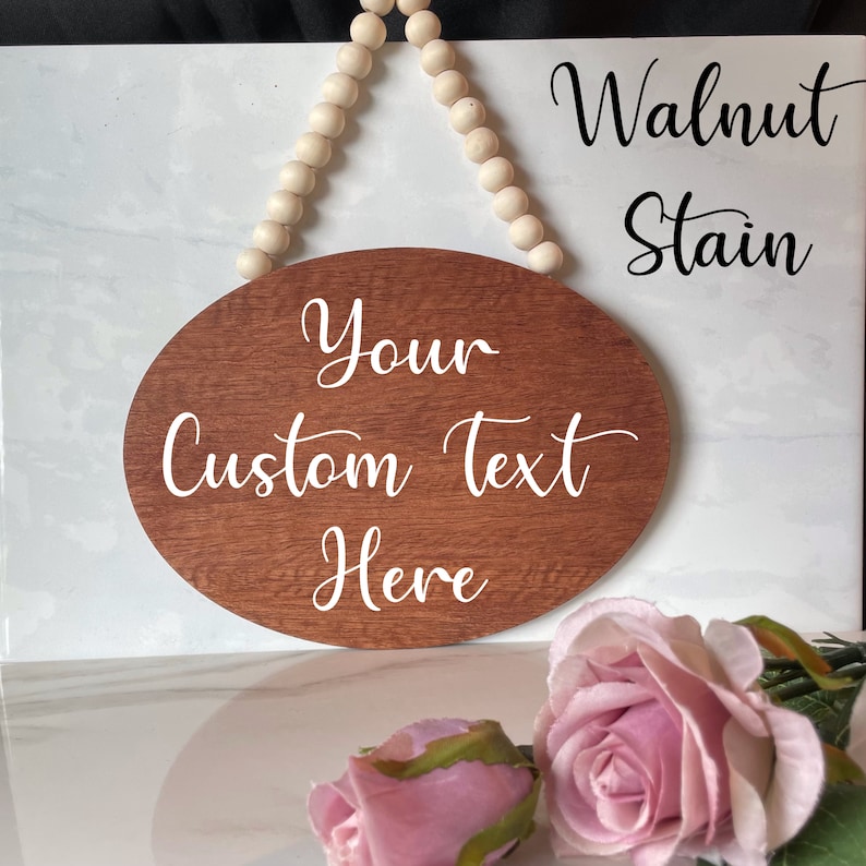 Pu&ograve; includere: Un cartello ovale in legno con una macchia di noce, appeso a una corda di perline bianche. Il cartello ha un testo bianco che dice "Your Custom Text Here".