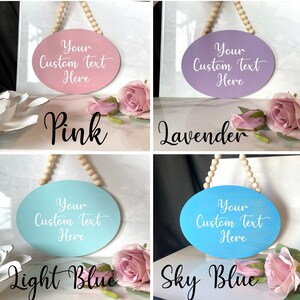 Pu&ograve; includere: Quattro cartelli ovali in legno con una corda di perline appesa in cima. I cartelli sono dipinti in rosa, lavanda, azzurro e celeste. Ogni cartello ha il testo "Your Custom Text Here" scritto in bianco.