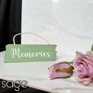 May include: A green wooden sign with the word "Memories" written in white. The sign is hanging from a white string and is sitting on a white marble surface. A pink rose is in the background.