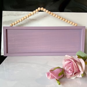 Peut inclure: Panneau rectangulaire en bois lavande avec un cintre en perles de bois. Le panneau a un design simple et encadr&eacute; et une finition vieillie. Une rose rose et un bouton sont au premier plan.