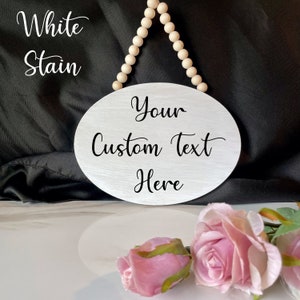 Pu&ograve; includere: Un cartello ovale in legno bianco con un appendiabiti a perline. Il cartello ha il testo "Your Custom Text Here" scritto in nero.