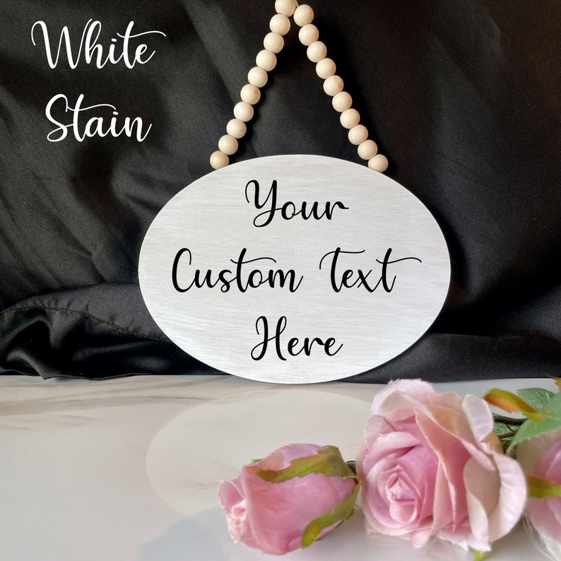 Pu&ograve; includere: Un cartello ovale in legno bianco con un appendiabiti a perline. Il cartello ha il testo "Your Custom Text Here" scritto in nero.