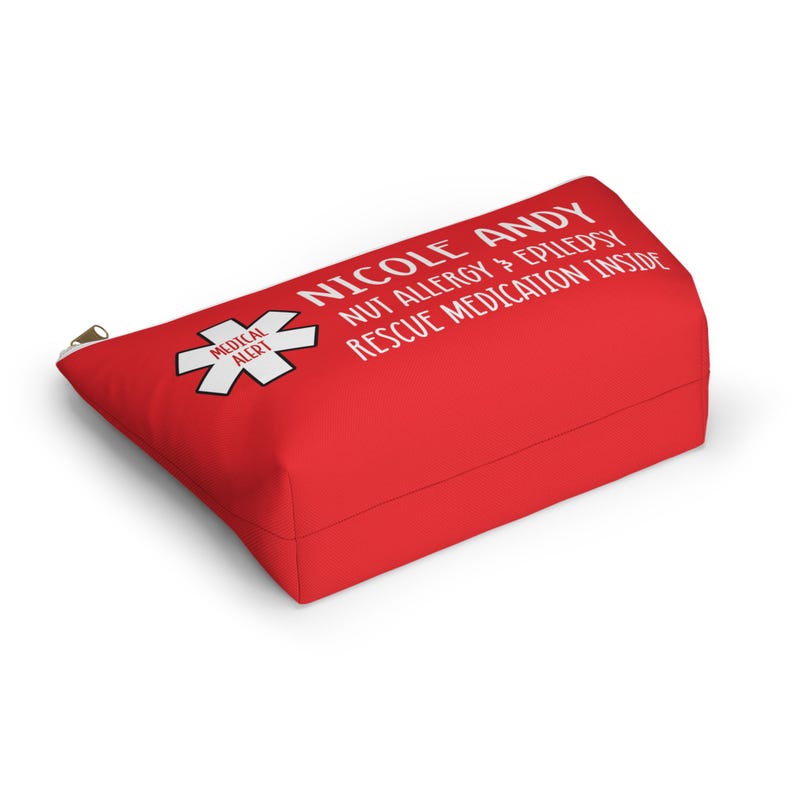 May include: Red zippered pouch with white text that reads "Nicole Andy Nut Allergy & Epilepsy Rescue Medication Inside". A red medical alert symbol with a white star is on the left side of the pouch.