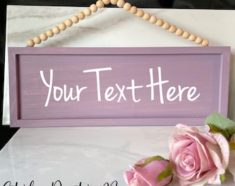 May include: A rectangular dusty mauve wooden sign featuring the white text Your Text Here. The sign is suspended by a string of natural light-colored wooden beads. Two artificial pink roses are positioned near the bottom right corner.