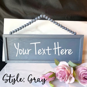 May include: Gray wooden sign with the text "Your Text Here" in white script. The sign is rectangular with a gray beaded hanger. The words "Style: Gray" are written in black script at the bottom of the sign. Pink roses are in the foreground.