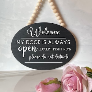 May include: Black oval sign with white text that reads "Welcome, My door is always open...except right now, please do not disturb". The sign is hanging from a wooden bead garland.