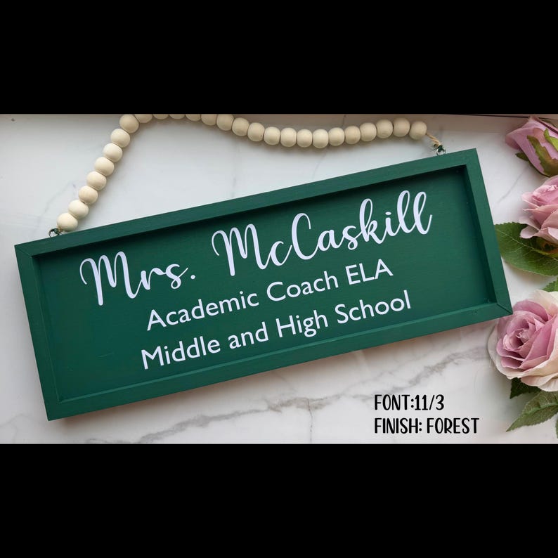 Peut inclure: Une enseigne rectangulaire vert for&ecirc;t avec des lettres blanches indiquant "Mrs. McCaskill, Academic Coach ELA, Middle and High School." L'enseigne est encadr&eacute;e et poss&egrave;de une cha&icirc;ne de perles pour l'accrocher. Le texte "FONT:11/3, FINISH: FOREST" est &eacute;galement visible.