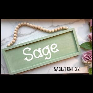 May include: A sage green wooden sign with the name "Sage" in white script. The sign is rectangular and has a beaded wooden garland above it. The text "SAGE/FONT 22" is below the name. The background is a marble-like surface.