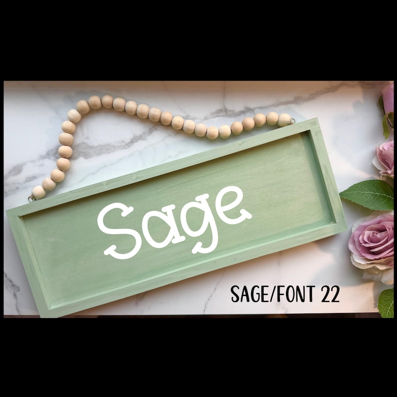 May include: A sage green wooden sign with the name "Sage" in white script. The sign is rectangular and has a beaded wooden garland above it. The text "SAGE/FONT 22" is below the name. The background is a marble-like surface.