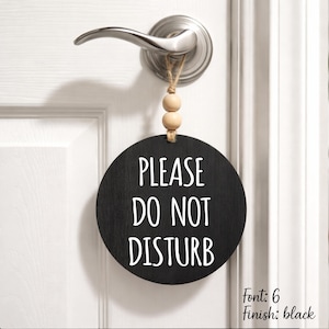 May include: A round black wooden door sign hanging from a silver door handle by a jute twine string with two light-colored wooden beads. The sign features the white text "PLEASE DO NOT DISTURB" in a clean, sans-serif font, centered on the surface.