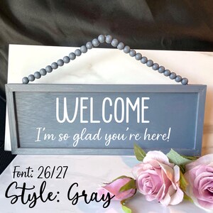Peut inclure: Panneau de bienvenue en bois gris avec le texte blanc "WELCOME I'm so glad you're here!". Le panneau est rectangulaire avec une poign&eacute;e grise perl&eacute;e. Des roses roses sont dans le coin inf&eacute;rieur droit. Le texte "Font: 26/27 Style: Gray" est en bas.