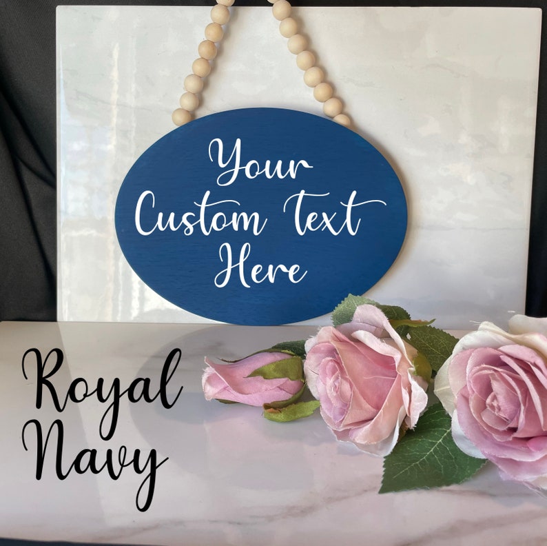 Pu&ograve; includere: Un cartello ovale in legno blu navy con un gancio in corda bianca. Il cartello dice "Your Custom Text Here" in lettere bianche in corsivo. Il cartello &egrave; appoggiato su una superficie di marmo bianco con fiori rosa.