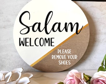 May include: Round wooden sign with a white and brown background. The sign says "Salam Welcome" in black cursive lettering on the white side and "Please Remove Your Shoes" in white lettering on the brown side.