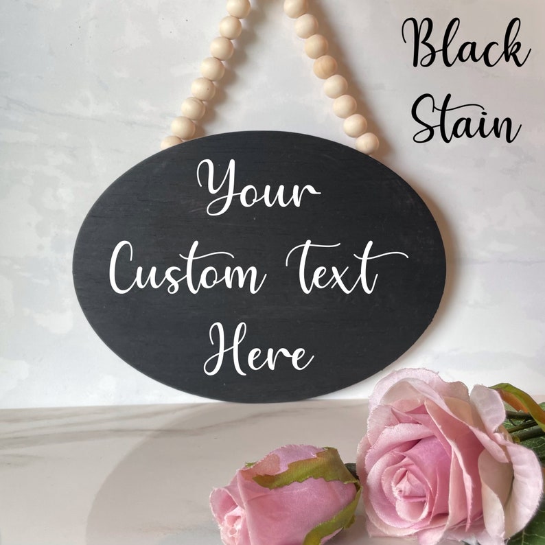 Pu&ograve; includere: Un cartello ovale in legno nero con testo bianco che recita "Your Custom Text Here". Il cartello &egrave; appeso a una corda di perline di legno. Due rose rosa sono in primo piano.