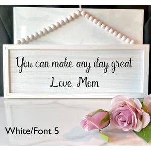 Peut inclure: Panneau en bois blanc avec l'inscription "You can make any day great Love, Mom" en noir. Le cadre est blanc et une guirlande de perles est suspendue. Des roses roses sont au premier plan.