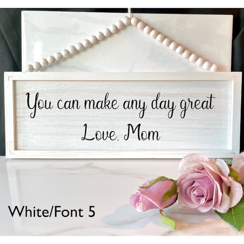 Peut inclure: Panneau en bois blanc avec l'inscription "You can make any day great Love, Mom" en noir. Le cadre est blanc et une guirlande de perles est suspendue. Des roses roses sont au premier plan.