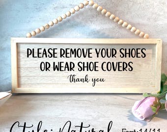 May include: A rectangular wooden sign with the text "PLEASE REMOVE YOUR SHOES OR WEAR SHOE COVERS Thank you." The sign is framed in light wood and is suspended by a string of wooden beads. The word "Natural" is written below the sign.