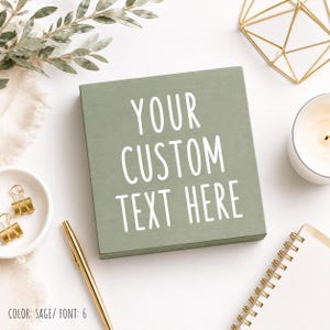May include: A square sage green wooden sign featuring the white text YOUR CUSTOM TEXT HERE in a clean sans-serif font. The sign is positioned centrally, surrounded by a gold pen, a small white bowl with gold clips, a candle, and a notebook.