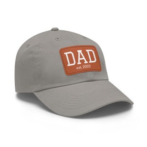 May include: A gray baseball cap with a brown leather patch that says "DAD est. 2023".
