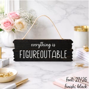 May include: A black wooden sign with the words "everything is FIGUREOUTABLE" in white lettering. The sign has a natural rope hanger. The background includes a vase of flowers and office supplies.