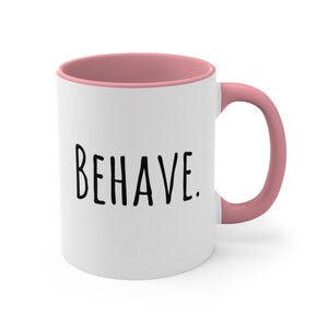 May include: White ceramic mug with a pink rim and handle. The mug has the word "BEHAVE." printed in black on the front.