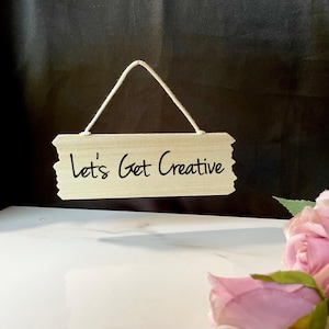 May include: A wooden sign with a white string hanging from the top. The sign has the text "Let's Get Creative" written in black.