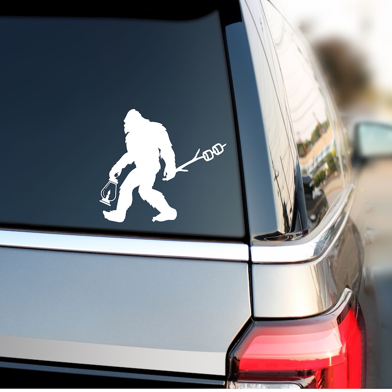 May include: White vinyl decal of a Bigfoot silhouette holding a lantern and a stick with three marshmallows on it. The decal is on the back window of a silver car.