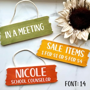 May include: Three wooden signs with white text on a white background. The signs are hanging from twine. The top sign is green and says "IN A MEETING". The middle sign is yellow and says "SALE ITEMS 1 FOR $1 OR 5 FOR $4". The bottom sign is orange and says "NICOLE SCHOOL COUNSELOR".