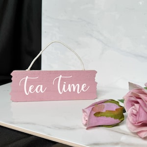 May include: A pink wooden sign with the text "tea time" in white cursive lettering. The sign has a string loop for hanging and is sitting on a white marble surface.