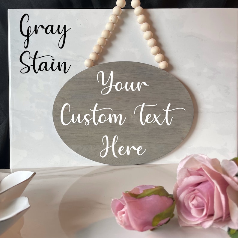 Pu&ograve; includere: Un cartello ovale in legno tinto di grigio con testo bianco che recita "Your Custom Text Here". Il cartello &egrave; appeso a una corda di perline bianche.