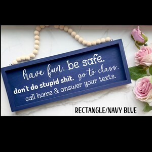 May include: A rectangular navy blue wooden sign with white text that reads: have fun. be safe. don't do stupid shit. go to class. call home & answer your texts. The sign features a light-colored wooden bead hanger attached to the top frame.