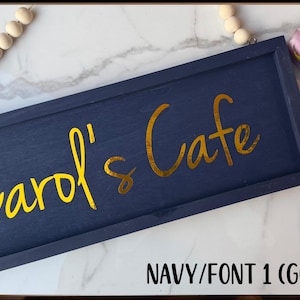 May include: Navy blue wooden sign with the text "Carol's Cafe" in gold script. The sign is rectangular and framed. A string of wooden beads hangs from the top. Pink roses are in the background.