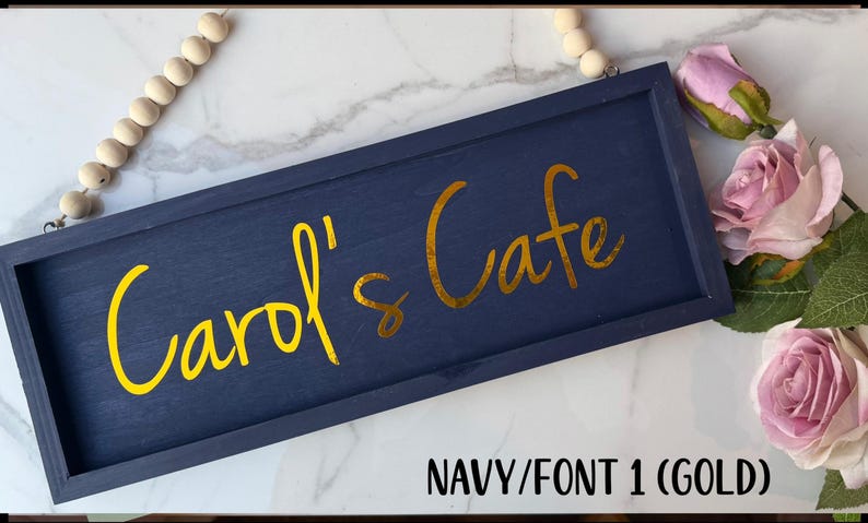 May include: Navy blue wooden sign with the text "Carol's Cafe" in gold script. The sign is rectangular and framed. A string of wooden beads hangs from the top. Pink roses are in the background.