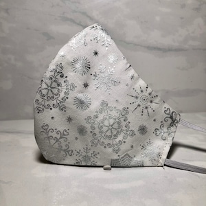 May include: White face mask with a silver snowflake pattern. The mask has elastic ear loops.